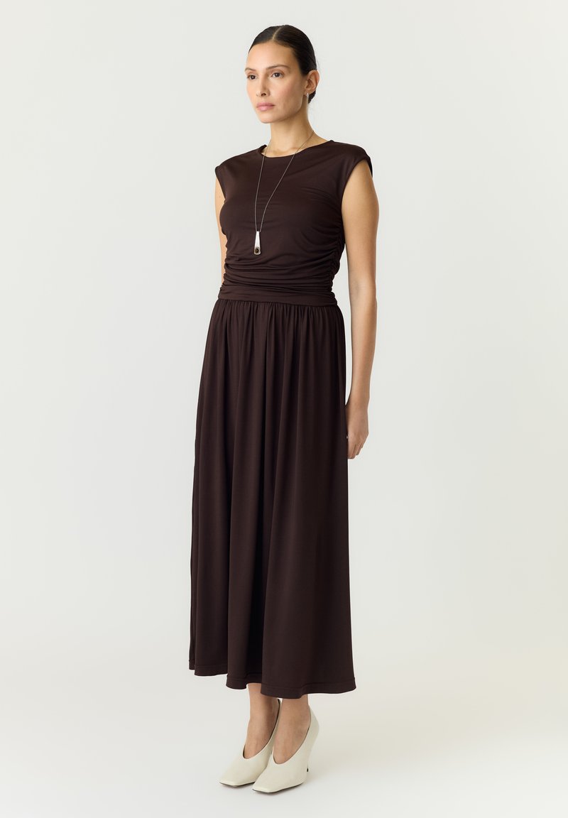 Femme avec les cheveux attachés portant une robe marron foncé sans manches et des chaussures blanches, debout devant un fond blanc uni.