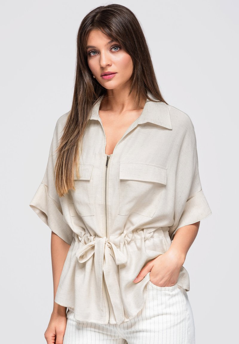 Jeune femme aux longs cheveux bruns portant une chemise beige clair zippée avec une ceinture à la taille et un pantalon blanc à fines rayures, main dans la poche, regardant devant elle.