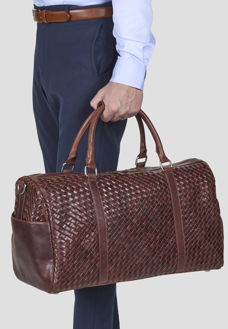 Hnědá tkaná kožená cestovní taška s pevným hnědým madlem, která má hladký kožený detail a zapínání na zip.