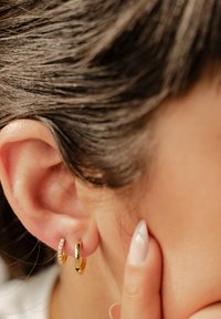 Pendientes de aro de oro con un acabado suave, uno de ellos adornado con piedras transparentes y brillantes. Vista cercana de la oreja y el dedo tocando la piel.