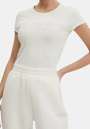 Donna che indossa un top bianco aderente a maniche corte e pantaloni della tuta bianchi elasticizzati a vita alta abbinati con tasche su uno sfondo semplice.