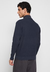 Donkerblauwe geribbelde trui met een opstaande kraag, voorzien van een gestructureerd breipatroon en een aansluitende pasvorm. Geen zichtbare hardware of accenten.