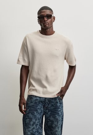 Homme portant un pull beige texturé à manches courtes et un jean bleu à motifs floraux, avec des lunettes de soleil noires, debout devant un fond clair uni.