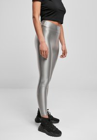 Hoog uitgesneden metallic zilveren legging met een gladde textuur en een naad aan de zijkant; gedragen met zwarte sportschoenen.