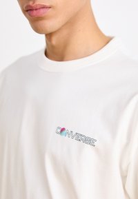 Hvid bomulds t-shirt med et farverigt "CONVERSE" logo på venstre brystområde, rund halsudskæring og korte ærmer.