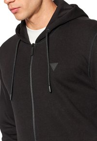 Schwarzer Kapuzenpullover mit Reißverschluss und Kordelzug an der Kapuze, gefertigt aus weichem Stoff, mit einem strukturierten Dreieck-Logo auf der linken Brust.