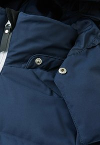 Dunkelblaue, gepolsterte Jacke im Detail, mit glattem Stoff, Druckknöpfen und hohem Kragen. Texturiertes Innenfutter ist teilweise sichtbar.