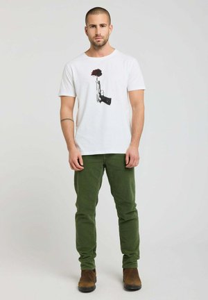 Homme aux cheveux courts portant un t-shirt blanc avec un motif de pistolet et de rose rouge, pantalon vert et bottes marron, se tenant devant un fond uni.