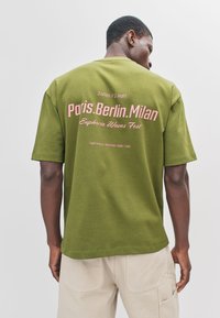 Donkergroen T-shirt met korte mouwen, met roze tekst op de achterkant, inclusief stadsnamen en festivalgegevens, gemaakt van zacht katoen.