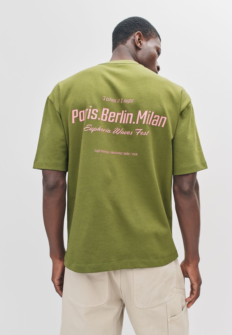 Donkergroen T-shirt met korte mouwen, met roze tekst op de achterkant, inclusief stadsnamen en festivalgegevens, gemaakt van zacht katoen.