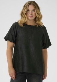 Schwarze Bluse mit strukturiertem, plissiertem Design. Verfügt über einen runden Ausschnitt und kurze Puffärmel. Kombiniert mit dunklen, eng anliegenden Jeans.