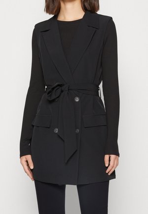 Kobieta ubrana w bezrękawnik w kolorze czarnym z paskiem, na czarnym, prążkowanym, długim rękawie, w połączeniu z czarnymi spodniami.