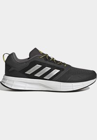 Zapatillas deportivas negras con parte superior de malla gris, franjas reflectantes plateadas y cordones con acentos amarillos. Suela blanca gruesa con agarre texturizado.