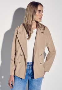 Beige double-breasted jacket met een puntige kraag, knoopaccenten en een relaxte pasvorm, gecombineerd met een witte top en blauwe jeans.
