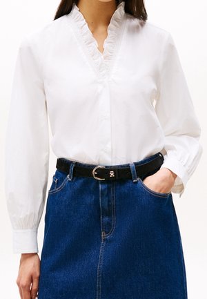 Femme portant un chemisier blanc à manches longues avec un col en V à volants et une jupe en denim bleue avec une ceinture noire, main dans la poche.