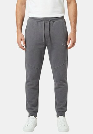 Homme portant un pantalon de jogging gris foncé avec une taille à cordon, des poches latérales et des baskets blanches sur un fond blanc.