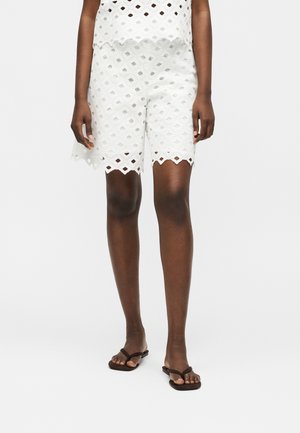Dolna część ciała osoby noszącej białe koronkowe szorty z ażurowym wzorem i czarne płaskie sandały na jednolitym jasnym tle.
