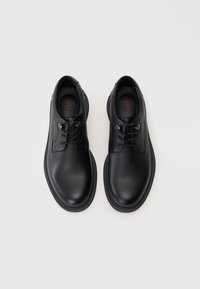 Chaussures noires en cuir avec un dessus texturé, bout rond, lacets plats et semelle en caoutchouc. La semelle intérieure présente un texte rouge.