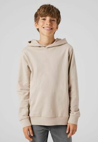 Långärmad beige hoodie i mjukt tyg med dragsko i huvan, ribbade ärmslut och en avspänd passform, kombinerad med grå jeans.