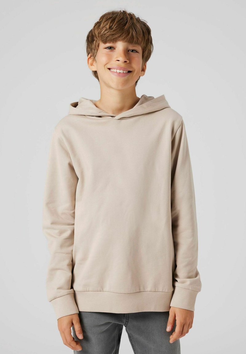 L�ångärmad beige hoodie i mjukt tyg med dragsko i huvan, ribbade ärmslut och en avspänd passform, kombinerad med grå jeans.