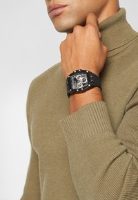 Montre rectangulaire noire avec un cadran clair, des accents argentés et un bracelet en silicone, portée au poignet, associée à un pull à col roulé beige.