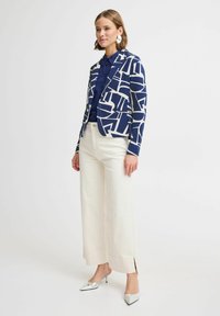 Blazer bleu marine et blanc à motifs avec des formes abstraites, associé à une chemise bleue et un pantalon large blanc, complété par des chaussures à talons argentées.
