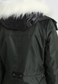 Giacca verde scuro con finitura lucida, dotata di cappuccio staccabile in pelliccia sintetica bianca, dettaglio nero sulle spalle e cintura in vita.