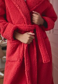 Red, fluffy bathrobe with a textured surface, wrap design, wide sleeves, and a matching belt. Features two front pockets.