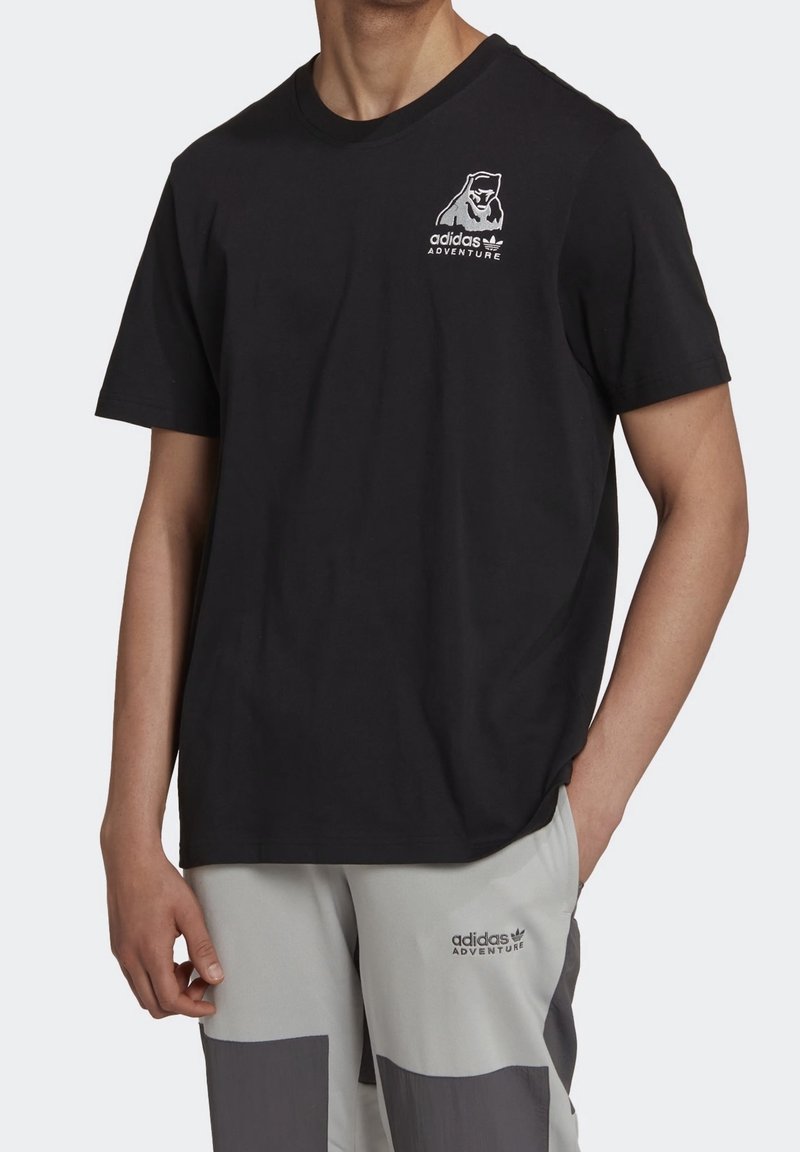 Man i svart Adidas Adventure T-shirt och ljusgrå byxor med mörkgrå detaljer, Adidas Adventure-logotyp på båda plaggen.