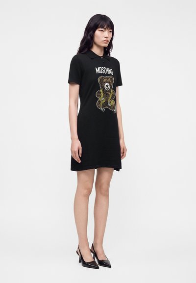Femme aux longs cheveux noirs portant une robe polo noire avec un ours en peluche et le texte "MOSCHINO", associée à des chaussures à talons hauts noires.