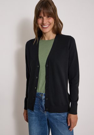 Femme souriante aux cheveux bruns portant un cardigan noir boutonné sur une chemise verte et un jean bleu sur un fond uni.
