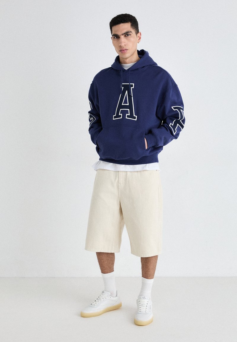 Marineblaues Hoodie mit weißem "A"-Logo und grafischen Ärmeln, kombiniert mit kremfarbigen Shorts und weißen Sneakers mit hellen Sohlen.