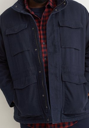 Homme portant une veste bleu marine avec quatre poches à rabats sur une chemise à carreaux rouge et noire.