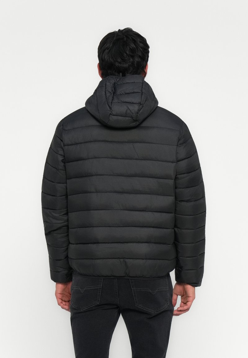 Man in een zwarte gewatteerde hoodie-jack, staand met zijn gezicht naar een witte muur, handen langs zijn zij, gekleed in zwarte jeans.