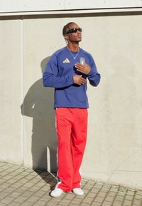 Modrá mikina s logom Adidas a talianskou vlajkou, doplnená červenými teplákmi s bielymi pruhmi a bielymi teniskami.