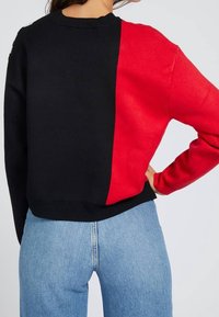 Pull noir et rouge avec un design color-block, col et poignets côtelés, offrant une coupe décontractée, associé à un jean bleu clair.