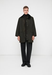 Olijfgroene jas met een kraag, drukknoopsluiting en zijzakken; gecombineerd met een zwarte coltrui en donkere jeans. Gladde, matte stof.