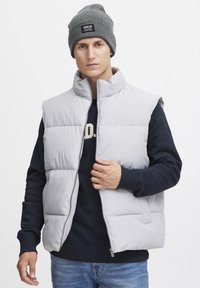 Light gray puffer vest with high collar, zip closure, and side pockets, worn over a dark sweater. Model also wears a gray knit beanie.