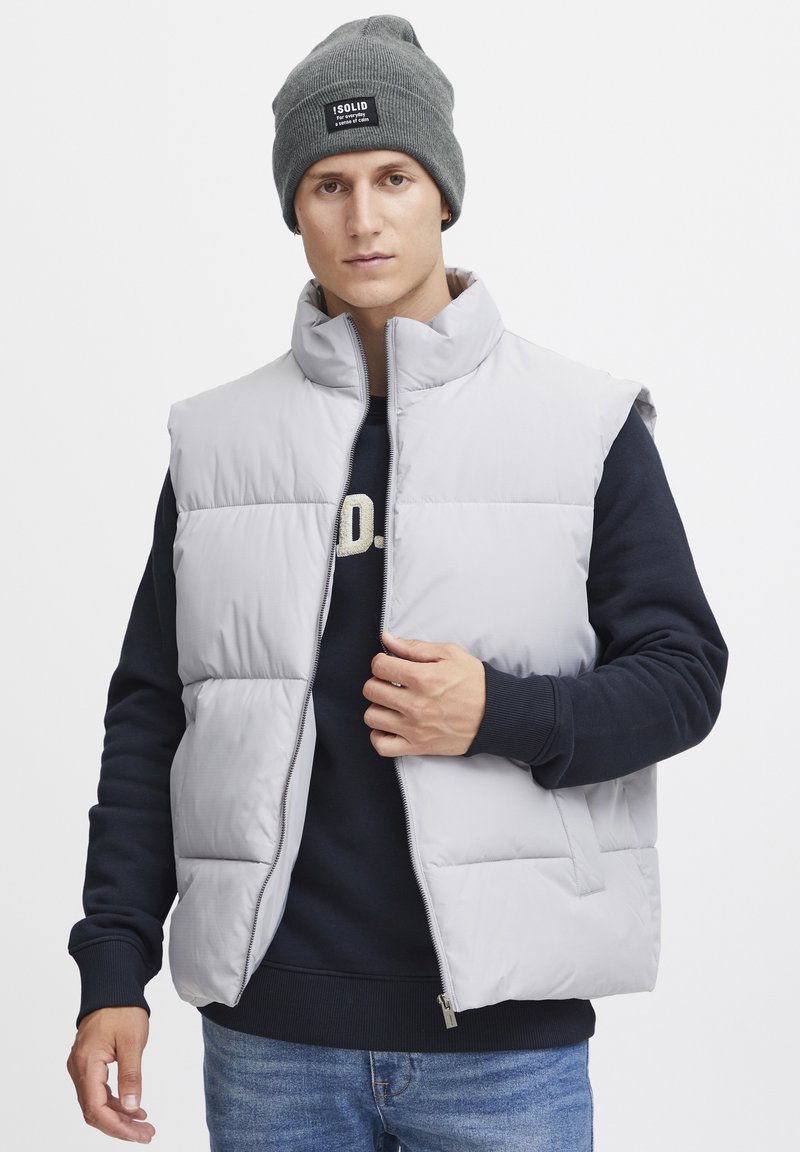 Light gray puffer vest with high collar, zip closure, and side pockets, worn over a dark sweater. Model also wears a gray knit beanie.
