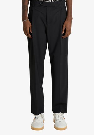 Uomo con pantaloni neri a gamba dritta e sneakers bianche con suola in gomma, in piedi davanti a uno sfondo semplice.