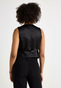 Gilet di raso nero con silhouette aderente, caratterizzato da un dettaglio a fiocco sul retro e scollatura alta. Indossato con pantaloni neri su misura.