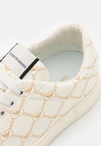 Baskets blanches avec chaîne beige et motif logo, lacets blancs, et une étiquette en tissu sur la languette avec le texte de la marque.