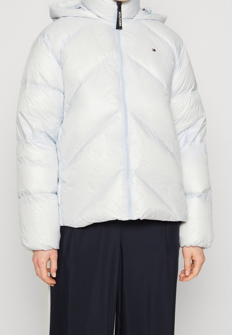 Veste matelassée bleu clair avec capuche, dotée d'un matelassage en chevrons, fermeture éclair à l'avant, poignets élastiques et un logo sur le côté gauche de la poitrine.