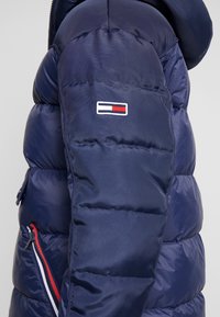 Granatowa pikowana kurtka puchowa z przeszyciami, z małym haftowanym logo oraz kieszenią na zamek z czerwono-białymi akcentami.