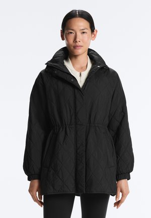 Frau mit dunklem Haar, die eine schwarze, gesteppte Jacke mit hohem Kragen, elastischen Bündchen und Taillenband trägt, vor einem schlichten weißen Hintergrund steht.