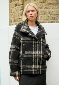 Veste en tartan noir et crème en tissu texturé, avec un col replié, des boutons-pression et des poches avant.