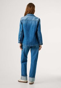 Denimjack met bijpassende spijkerbroek in een lichte naar donkere blauwe gradient. Voorzien van knoopmanchetten, zichtbare stiksels en opgerolde manchetten.