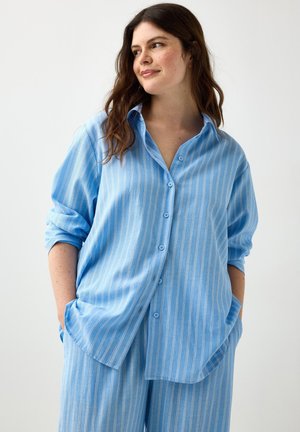 Donna con capelli castani lunghi che indossa una camicia azzurro chiaro a righe verticali ampia e pantaloni abbinati, mani in tasca, guarda di lato.