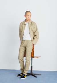 Beige jakke og matchende bukser laget av lett materiale, kombinert med en hvit t-skjorte og svarte sko med gule striper.