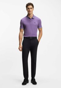 Polo viola con colletto a righe nere e bianche a contrasto, abbinata a pantaloni neri e scarpe nere, in piedi contro uno sfondo semplice.